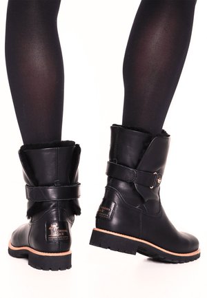 Bottes en cuir noir mi-mollet avec doublure en fourrure, sangles réglables et semelles épaisses portées avec des collants noirs sur fond blanc.