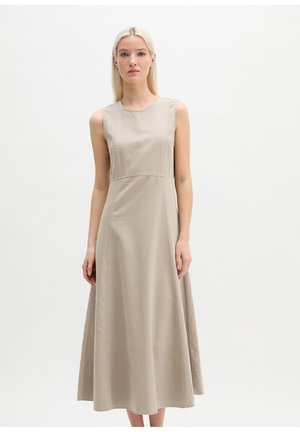 Młoda kobieta z długimi blond włosami, ubrana w bezrękawnikową beżową sukienkę midi, stojąca na tle jednolitego białego tła.