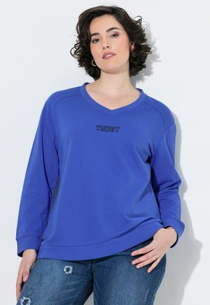 Donna con capelli corti e ricci che indossa una felpa blu "Midnight Muse" e jeans blu strappati, in piedi davanti a uno sfondo bianco.