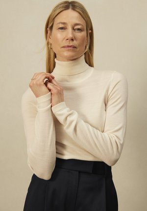 Crème coltrui in een lichte brei. Heeft aansluitende mouwen en een gladde textuur, gecombineerd met hoge, donkere broek.