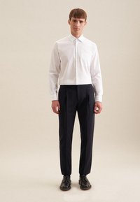 Witte cropped shirt met knoopsluiting, lange mouwen en één borstzak, gecombineerd met marineblauwe geplooide broek en zwarte veterschoenen.