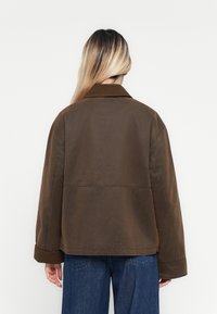 Žena s dlouhými blond vlasy, oblečená v volné hnědé bundě a modrých džínech, stojící zády k jinak bílé pozadí.