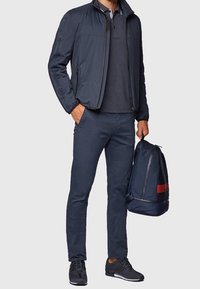 Marinblå lättviktjacka, matchande polo och byxor, parat med en mörk ryggsäck med röda detaljer. Skorna är mörka med texturerade ytor.