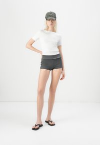 Hvit tettsittende t-skjorte, grå ribbede shorts, grå caps med svarte detaljer, svarte badesandaler, står mot en minimal hvit bakgrunn.