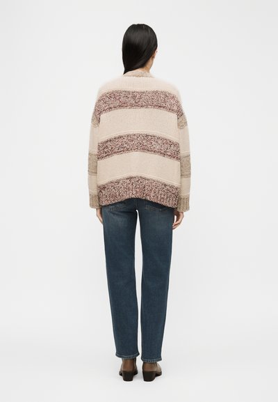 Γυναίκα με μακριά σκούρα μαλλιά φοράει υπερμεγέθη μπεζ πλεκτό πουλόβερ με μπορντό και καφέ ρίγες, στενό μπλε τζιν και καφέ παπούτσια με χαμηλό τακούνι, όρθια.