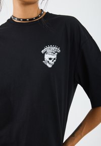 T-shirt en coton noir à coupe oversize, avec un graphisme de tête de mort blanche et le texte "Accro aux ramen" sur la zone gauche de la poitrine.