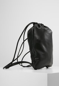 Sac à dos en cuir noir à cordon avec des bretelles fines, présenté en position verticale sur un socle blanc sur un fond gris clair.