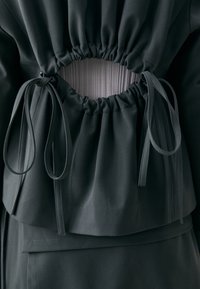 Dark green garment featuring a gathered back with a circular cutout, adjustable ties, and textured ribbed fabric underneath.
