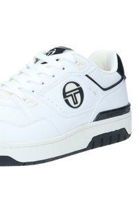 Sneaker bianco con sezioni perforate, dettagli neri e un logo rotondo. Presenta una suola piatta con un motivo testurizzato per la presa.