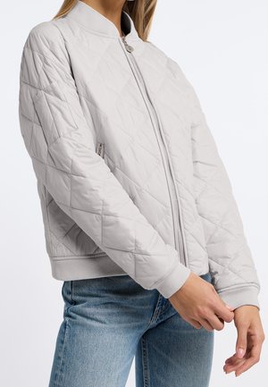 Femme portant une veste bomber matelassée gris clair avec fermeture éclair devant et un jean bleu, mains ajustant le poignet de la manche.