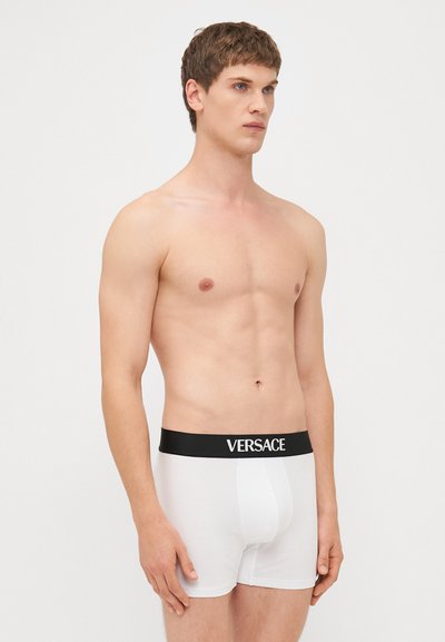 Fehér pamut bokseralsó fekete elasztikus derékpánttal, amelyen az elülső oldalon feltűnően van elhelyezve a "VERSACE" logó.