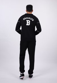 Black sweatshirt with white "BROOKLYN" text and "B" logo on the back. Paired with black sweatpants and black shoes with white soles.