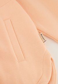 Sudadera melocotón con una textura suave, que cuenta con un bolsillo lateral y una pequeña etiqueta de marca, destacando un diseño simple y funcional.