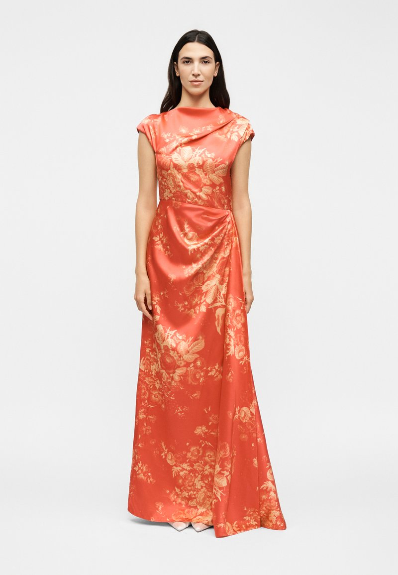 Woman wearing a floor-length, sleeveless coral satin dress with gold floral patterns, standing straight against a plain background.