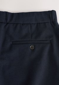 Dunkelblaue Hose mit elastischem Bund und einer einzelnen Gesäßtasche, die mit einem schwarzen Knopf gesichert ist.