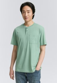 Tricou scurt cu mânecă, de culoare verde mentă, cu decolteu rotund și o plachetă cu trei butoane, având un buzunar pe pieptul stâng și fabricat din bumbac moale.