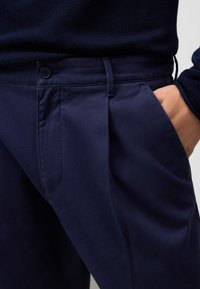 Marineblauwe broek van textuurstof met een knoopsluiting, plooien en zijzakken, met een close-up van het taillebandgedeelte.