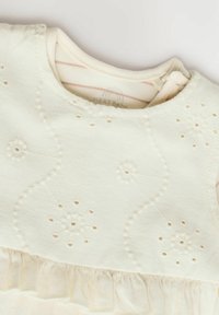 Robe en coton crème ornée de motifs floraux brodés, avec un col rond et une volantruffle froncée le long de l'ourlet. Tissu légèrement texturé.