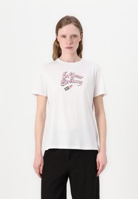Tricou alb din bumbac cu text pastel roz "În visele tale" și un grafic ilustrativ cu un ruj pe față. Mâneci scurte, gât rotund.