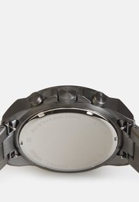Uhrende aus Edelstahl mit einer glatten, reflektierenden Oberfläche. Zwei Drücker und eine strukturierte Krone sind an der Seite sichtbar. Schwarzes Metallarmband.