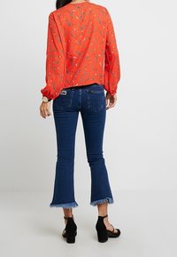 Röd mönstrad blus med långa ärmar, i kombination med blå utsvängda jeans med fransade fållar och en synlig varumärkesetikett. Svarta högklackade skor.