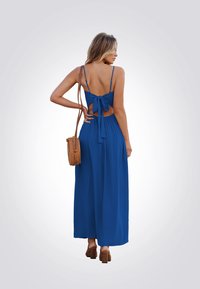 Vestido maxi azul con tirantes finos, espalda al aire y detalle de lazo. Un bolso marrón texturizado y zapatos de tacón ancho complementan el conjunto.