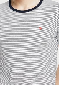 Close-up van een persoon die een wit T-shirt draagt met dunne zwarte strepen en een klein rood naaimachine-logo op de borst.