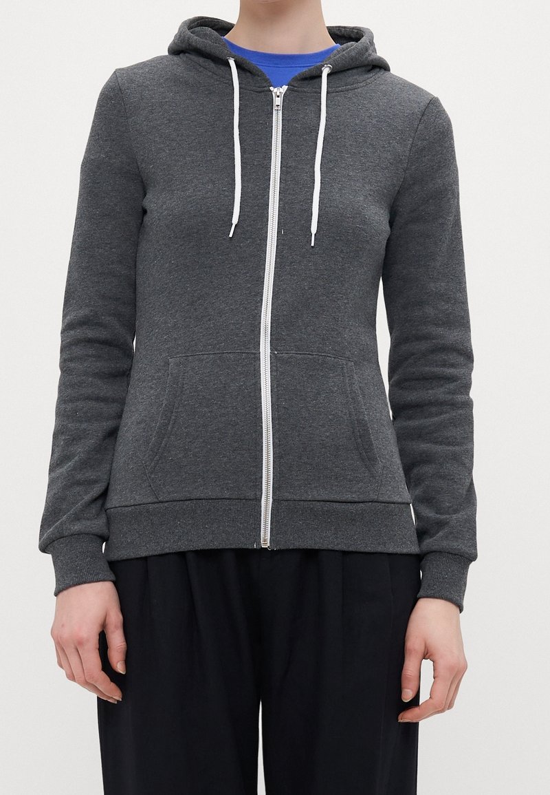Personne portant un sweat à capuche zippé gris foncé avec des cordons blancs sur une chemise bleue et un pantalon noir, debout devant un fond clair uni.