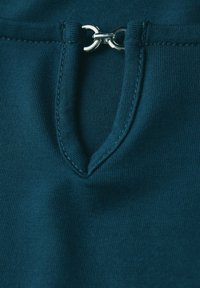 Teal stof met een gladde textuur heeft een kleine v-vormige opening bij de hals, bevestigd door een zilveren metalen lus en stiksel details.