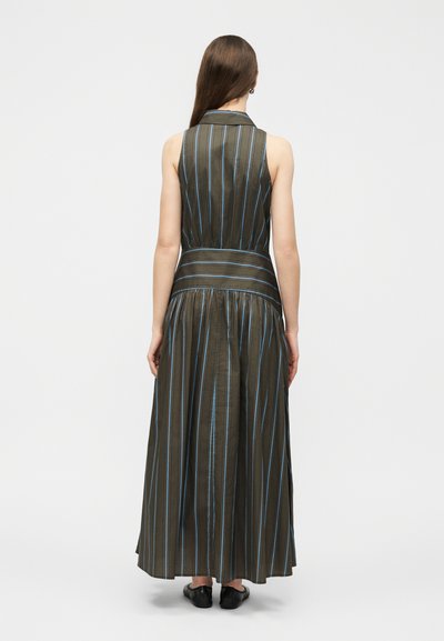 Γυναίκα που φοράει αμάνικο, μακρύ ελιά φόρεμα με γιακά και κάθετες μπλε ρίγες, στεκόμενη πλάτη σε απλό ανοιχτό φόντο.