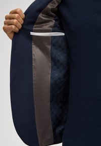 Mano che tiene aperto un blazer blu navy con fodera scura a motivi e taschino interno al petto rifinito in bianco visibile.