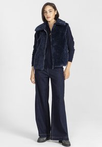 Kobieta stoi, ubrana w granatowy kamizelkę z sztucznego futra na długim rękawie, szerokie ciemnojeansowe spodnie i szare buty na białym tle.