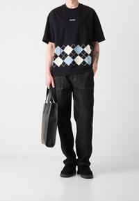 Schwarzes Kurzarm-T-Shirt mit einem diamantförmigen Muster in Blau und Weiß am Saum, kombiniert mit dunklen Jeans und einer schwarzen Tasche.