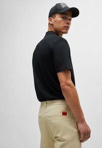 Black short-sleeve polo shirt paired with light beige trousers featuring a red "HUGO" tag on the back, presented from a slightly turned perspective.