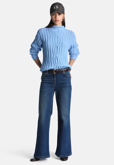 Femme portant un pull en maille torsadée bleu clair, un jean évasé bleu foncé, une ceinture noire, des chaussures noires et une casquette noire texturée avec des lettres blanches.