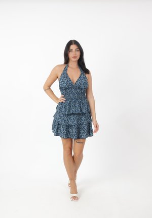 Vrouw met lang donker haar die een blauwe bloemenprint halterjurk en witte open teenhakken draagt, staand tegen een witte achtergrond.