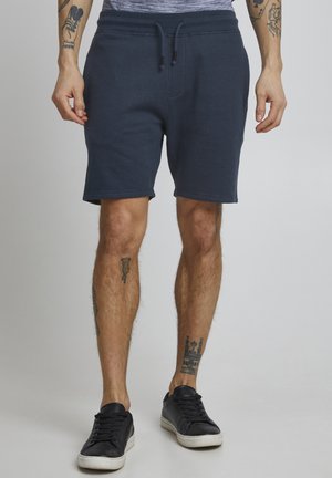 Mørkeblå bomuldsshorts med en struktureret overflade, elastisk talje og justerbar snor. Båret med sorte sneakers og synlige tatoveringer.