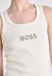 Witte geribde tanktop met een gestructureerde logopatch met de tekst "BOSS" in zilveren strassstenen. Ronde halslijn en getailleerd ontwerp.