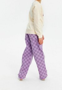 Luźne, fioletowe dżinsy w kratkę z postrzępionymi nogawkami, zestawione z kremową bluzą z graficznym wzorem, pokazane z boku.