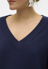 Navy V-Ausschnitt-Pullover mit geripptem Kragen und weicher Textur, dargestellt mit einem teilweise sichtbaren Nacken und Ohrstecker eines Models.
