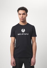 Juodas medvilninis marškinėlis su baltu logotipu ir užrašu "BELSTAFF" priekyje. Trumpomis rankovėmis ir klasikiniu apykaklės dizainu.