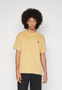 Camiseta de manga corta color mostaza con cuello redondo, que presenta un pequeño parche de logo cuadrado en el lado izquierdo del pecho, fabricada con suave tejido de algodón.
