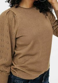 Pull en tricot marron avec un col rond, des manches bouffantes et de subtils détails ajourés sur les bras, assorti à un jean foncé.