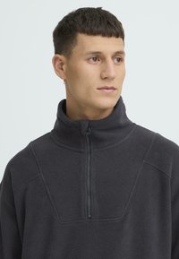 Tumeda halli fliisist pullover poollukuga kaelusega, tekstuurse kangaga ja õmblustega paneelid õlgadel ja rinnal.