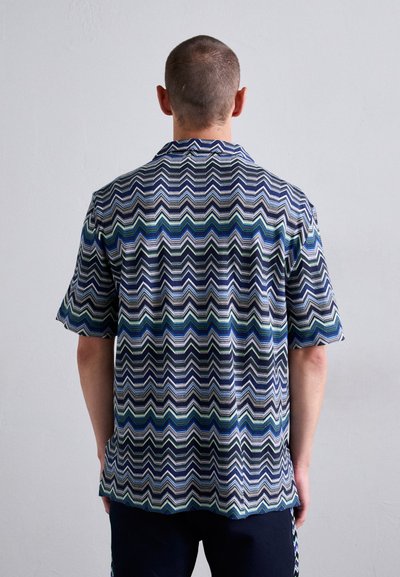 Uomo con capelli corti che indossa una camicia a maniche corte con un motivo a zigzag nei colori blu, verde, bianco e nero, in piedi di fronte a un muro chiaro e semplice.