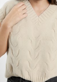 Maglione beige a treccia con scollo a V e orlo a coste. Tessuto morbido con sottili accenti di lucentezza in tutto il materiale.