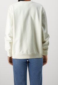 Persona che indossa una felpa semplice color crema e jeans di denim blu, vista di spalle su uno sfondo neutro.