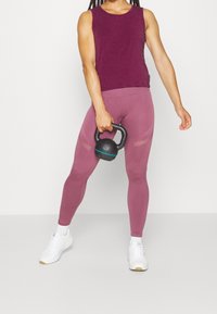 Lila ärmlös topp och åtsittande rosa leggings med nätdetaljer, håller en svart kettlebell, kombinerat med vita sportskor mot en neutral bakgrund.