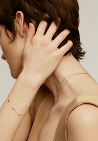 Joyería de oro con cadenas delicadas, una de ellas con pequeñas piedras de colores, llevada en la muñeca y el cuello. Blusa de tela suave con tirante ancho.