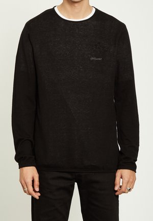 Mann trägt ein schwarzes Langarmshirt mit kleinem Brustlogo und weißem Kragenabschluss, kombiniert mit schwarzer Hose und silbernen Ringen an den Fingern.